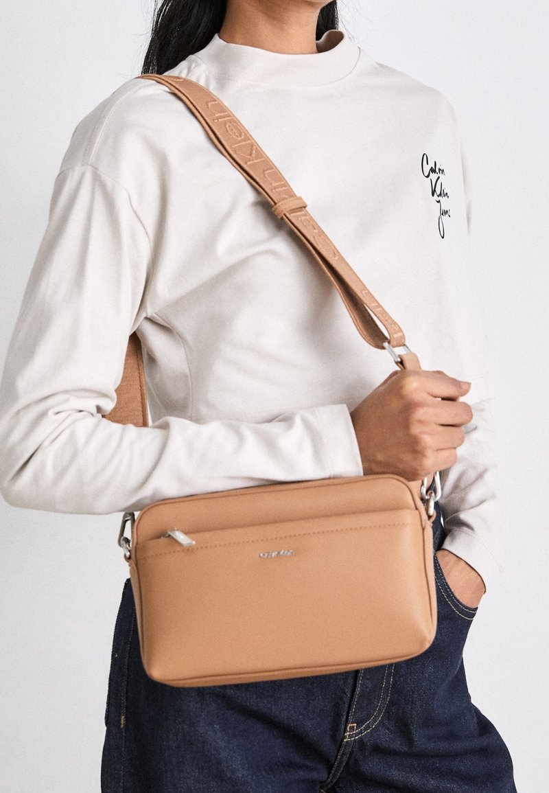 Femme en chemise blanche à manches longues et pantalon foncé portant un sac bandoulière beige avec une sangle réglable sur l'épaule.