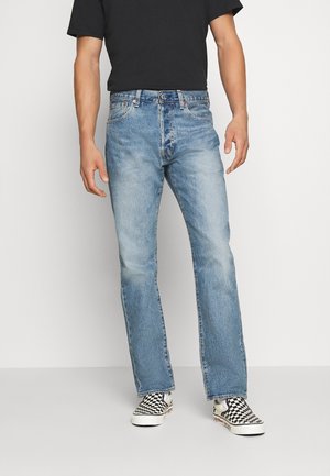 Mann trägt hellblaue Jeans mit geradem Bein, schwarzes T-Shirt und schwarz-weiß karierte Slip-On-Sneaker, steht vor einem einfarbigen Hintergrund.