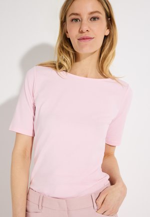 Woman with blonde hair wearing a light pink short-sleeve top and matching pink pants, posing against a plain white background.
