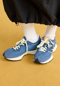 Baskets en denim bleu avec lacets blancs, tige texturée, semelle en caoutchouc et accents gris clair. Associées à des chaussettes blanches côtelées.