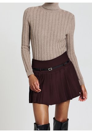 Femme portant un pull à col roulé beige en maille torsadée, une jupe plissée marron foncé avec une ceinture noire, et des bottes noires montantes sur fond blanc.