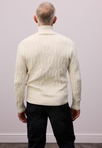Suéter color crema de punto trenzado con cuello alto, puños y dobladillo acanalados; combinado con jeans de mezclilla oscuros; vista trasera.