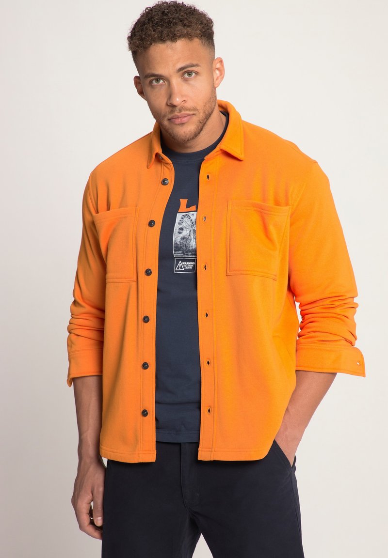 Camisa de franela naranja con cierre de botón, dos bolsillos en el pecho y mangas largas, superpuesta sobre una camiseta navy con estampado gráfico.