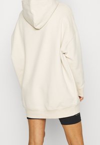 Sudadera de gran tamaño en color crema con capucha ajustable, puños de canalé y un tejido texturizado. Lleva puesta sobre pantalones cortos negros, mostrando un ajuste relajado.
