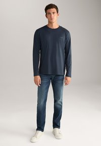 Marineblaues Langarmshirt mit Rundhalsausschnitt, leichtes Material, kombiniert mit ausgewaschenen blauen Jeans und weißen Sneakers, vor einem schlichten Hintergrund stehend.