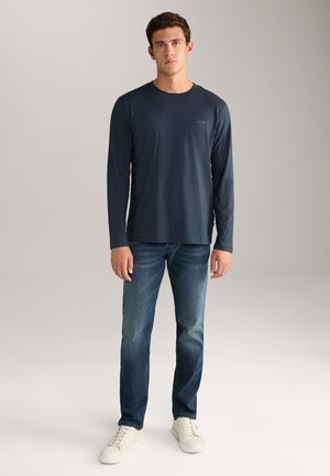 Navy long-sleeve shirt with a round neck, lightweight fabric, paired with faded blue jeans and white sneakers, standing on a plain background.