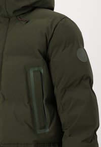 Grüne Pufferjacke mit Kapuze, strukturiertem Material, einem Patch-Logo auf dem Ärmel und einem kontrastierenden Taschenakzent.