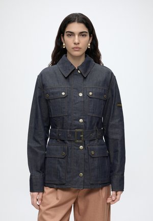 Vrouw met lang donker haar draagt een donker denim jack met riem en knoopzakken en beige broek tegen een effen lichte achtergrond.