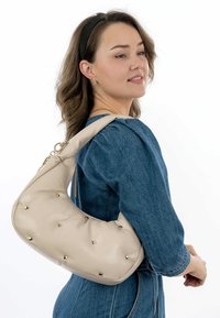 Borsa a tracolla in pelle beige con un design curvo, decorata con borchie dorate, indossata sulla spalla. Sfondo di tessuto semplice.