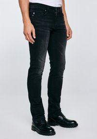 Férfi fekete slim-fit farmerben és fekete bőr fűzős cipőben, világos egyszínű háttér előtt állva.