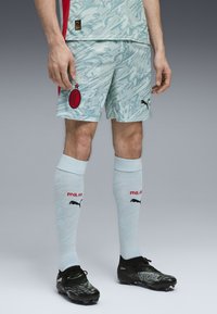 Completo da calcio azzurro chiaro con design astratto, dettagli rossi e logo dell'AC Milan. Scarponi neri con dettagli verdi e calze a compressione bianche.