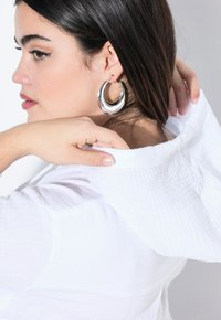 Garment bianco con cappuccio, texturizzato e dalla vestibilità rilassata, indossato da una persona con una mano poggiata sul collo. Sono visibili grandi orecchini a cerchio d'argento.