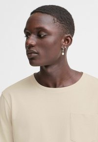 Beige színű póló, kerek nyakkivágással és zsebbetéttel. A modellen rövid haj van, és egy kis, gyöngy kiegészítőt visel a fülében.