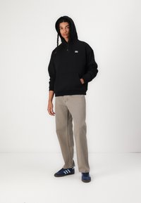 Schwarzer Kapuzenpullover mit einer Fronttasche, kombiniert mit hellbeigen, geraden Hosen und blauen Schuhen mit drei Streifen. Einfaches Design.