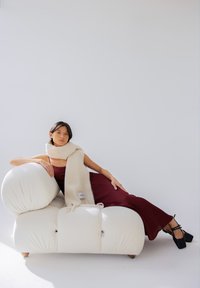 Crèmekleurige, pluche bank met ronde vormen en zichtbare naden, gecombineerd met een lange, zachte, beige sjaal met een label. Model in een kastanjebruine jurk.