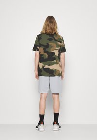 Maskáčové tričko ve odstínech zelené a hnědé, spárované se šedými šortkami. Model má na sobě černé tenisky s oranžovými akcenty a černé ponožky.