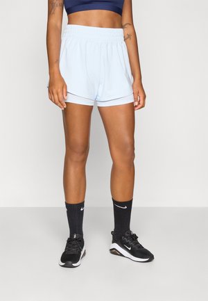 Žena ve světle modrých sportovních šortkách, černém sportovním topu, černých ponožkách značky Nike a černých běžeckých botách Nike, stojící na jednobarevném pozadí.