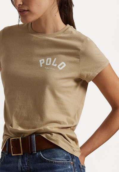 Beżowa bawełniana koszulka z okrągłym dekoltem, z białym nadrukiem logo "POLO" i krótkimi rękawami, zestawiona z niebieskimi dżinsami i brązowym paskiem.