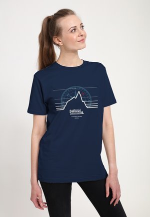 Kobieta z kucykiem, ubrana w granatowy T-shirt z motywem konturu góry, kompasu oraz napisem "Durham Mountaineering", stojąca na białym tle.