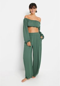 Zelena kratka majica z off-the-shoulder izrezom in dolgimi rokavi, v kombinaciji z ohlapnimi hlačami širokega kroja. Iz lahkotnega materiala z gladko teksturo.