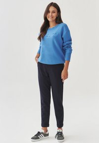 Felpa blu con testo in rilievo "BALANCE", abbinata a pantaloni tapered blu navy e sneakers grigie con lacci neri e dettagli bianchi.