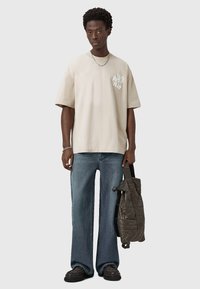 Man wearing oversized beige T-shirt, loose blue jeans, dark shoes, silver chain necklace and bracelet, holding a black quilted jacket bag.