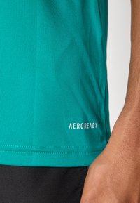 Turkis ermeløs treningsskjorte. Har teksturert stoff med en flat kant. Aeroready-logo i hvitt på nedre venstre side.