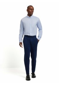 Helles blaues Hemd mit Knopfverschluss, kombiniert mit navyfarbener, taillierter Hose und schwarzen Dressschuhen. Das Hemd hat einen klassischen Kragen und lange Ärmel.