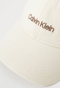 Hvit teksturert baseballcaps med en brodert brun "Calvin Klein" logo på fronten. Den har en buet skjerm og en strukturert design.