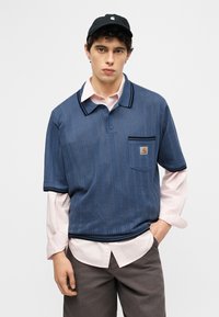 Νεαρός άντρας με μαύρο καπέλο, μπλε πλεκτό πόλο με κοντά μανίκια πάνω από ροζ ανοιχτό πουκάμισο και σκούρο παντελόνι, στέκεται με το ένα χέρι στην τσέπη.