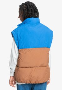 Puffer vest with a blue upper and brown lower section. Features a high collar, sleeveless design, and textured fabric.