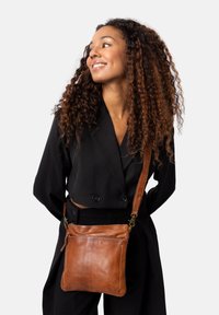 Borsa a tracolla in pelle marrone con una superficie liscia, manico robusto e tasca con cerniera, indossata su un outfit nero.
