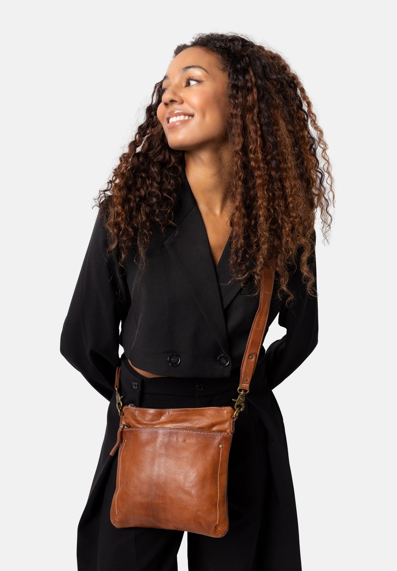 Borsa a tracolla in pelle marrone con una superficie liscia, manico robusto e tasca con cerniera, indossata su un outfit nero.