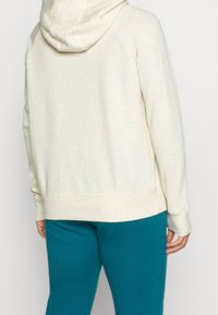 Felpa leggera color crema con finitura testurizzata, dotata di cappuccio con cordino e polsini a costine, abbinata a pantaloni teal. Design semplice e casual.