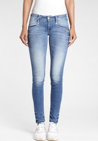 Slim-fit světle modré džíny, které má na sobě osoba stojící vzpřímeně s bílými teniskami a bílým topem, na jednobarevném světlém pozadí.