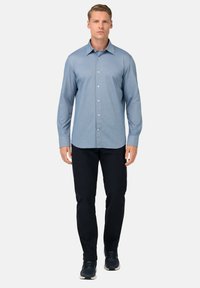 Camicia azzurra a maniche lunghe con colletto alla francese, abbinata a pantaloni scuri e sneakers, su uno sfondo bianco semplice.