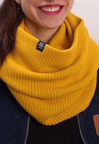 Yellow knitted infinity scarf with a ribbed texture, featuring a small black logo tag. Worn over a dark sweater and a quilted blue jacket.