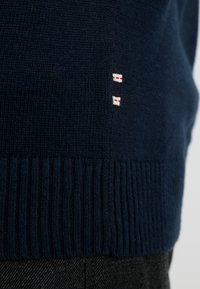 Zbliżenie na rąbek ciemnogranatowego dzianinowego swetra z prążkowanym wykończeniem i małą pionową naszywką w kolorach czerwonym, białym i granatowym, umieszczoną powyżej talii czarnych spodni.