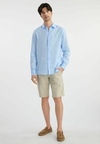 Jasnoniebieska koszula z długim rękawem i kołnierzykiem zapinanym na guziki, w połączeniu z jasnobeżowymi szortami i brązowymi butami wsuwanymi. Gładki materiał, swobodny krój.