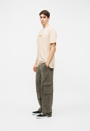 Νέος άνδρας που στέκεται πλαγίως φορώντας μια μπεζ μπλούζα με κείμενο, χακί παντελόνια cargo και μαύρα αθλητικά παπούτσια, με φόντο ένα απλό λευκό τοίχο.