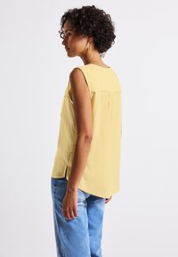 Blouse sans manches jaune avec détail de plis au dos, en tissu doux et fentes latérales, associée à un jean bleu clair à coupe décontractée.