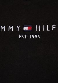 T-shirt in cotone nero con testo ricamato bianco "TOMMY HILFIGER" e logo rettangolare in rosso, bianco e blu; data di fondazione "EST. 1985."