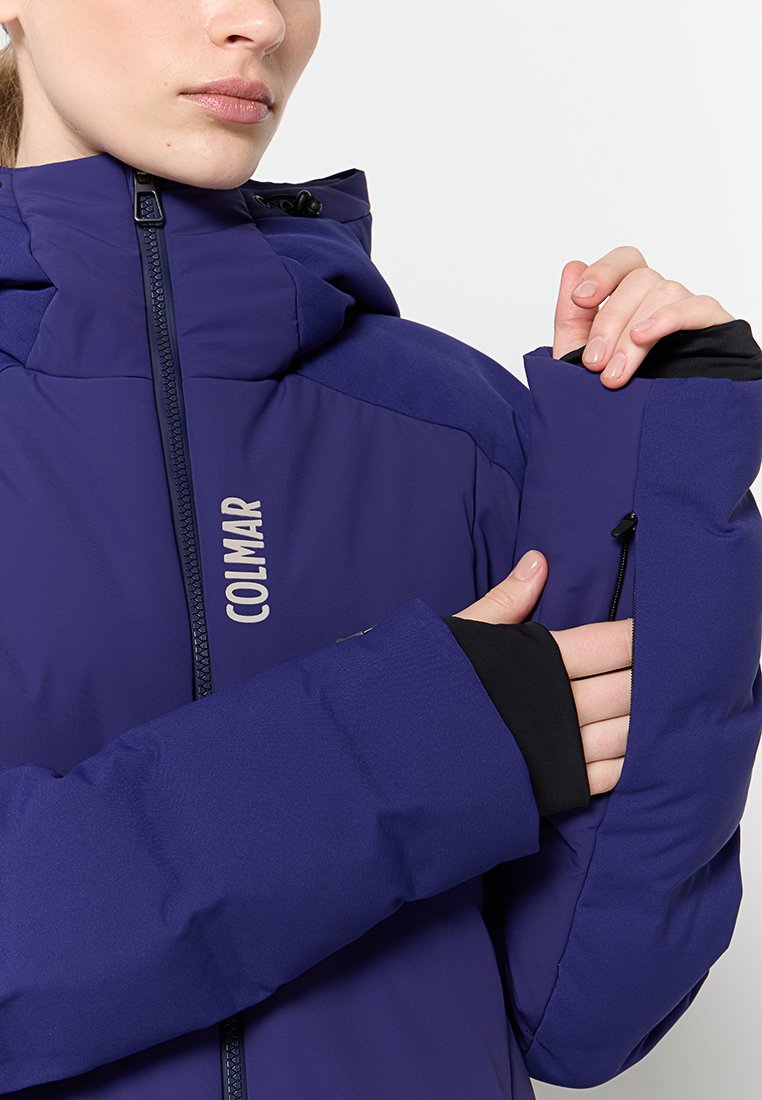Giacca isolante viola con colletto alto, dettagli neri e tasca laterale con zip. Il marchio "COLMAR" è visibile sul fronte.