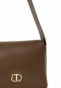 Borsa a spalla in pelle marrone con una texture liscia, chiusura con zip e un elemento in metallo oro con la lettera "T."