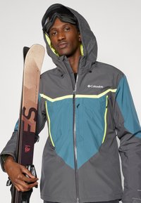 Casaco de esqui cinza e verde-água com detalhes amarelos, com fecho frontal e capuz. Modelo segura esquis e usa óculos de proteção, apresentando um design elegante.