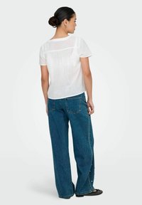Weißes kurzärmliges Blusenshirt mit strukturiertem Rücken, kombiniert mit hochgeschnittenen dunkelblauen weit geschnittenen Jeans. Schwarze flache Schuhe mit einem geflochtenen Muster.