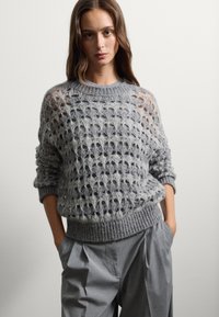 Maglione grigio spesso a maglia con un motivo a punto aperto, orlo a coste largo e polsini arrotolati, abbinato a pantaloni grigi plissettati.