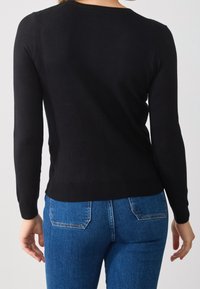 Mujer vestida con una blusa negra ajustada de manga larga y jeans de mezclilla azules, vista desde atrás sobre un fondo claro y liso.
