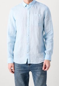 Camicia azzurra chiara con bottoni, realizzata in tessuto testurizzato, con una tasca sul petto, maniche lunghe e orlo curvo. Indossata con jeans.