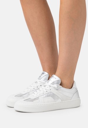 Jambes portant des baskets blanches basses avec des accents argentés et des détails perforés, posées sur un fond clair uni.